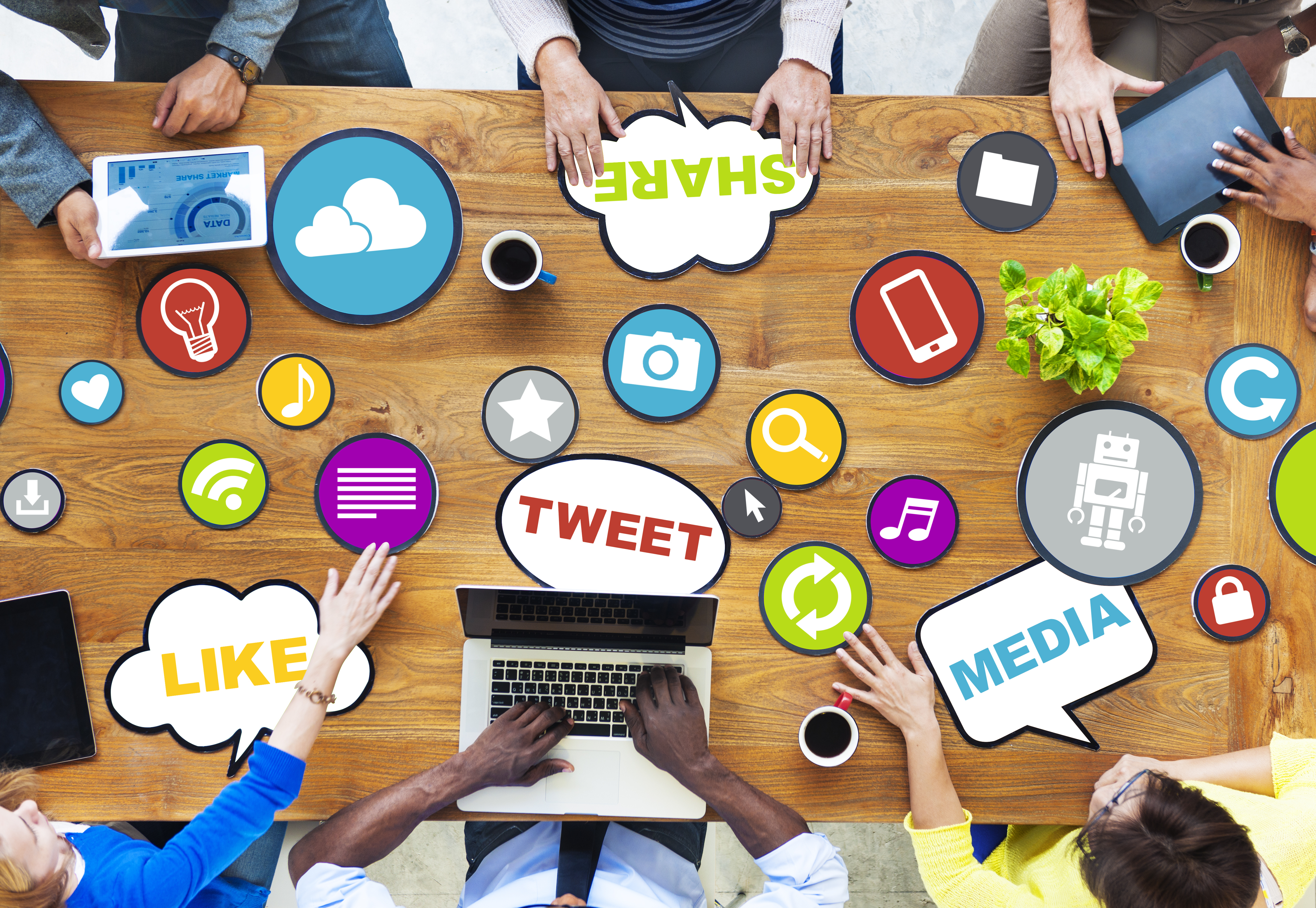 This picture shows a group of people gathered at a table with their computers. On the table are different cutouts of what appears to be paper with different media tags: share, tweet, nedia, likes. There are also more cutouts with symbols used for applications: download, music note, navigation bars, etc...